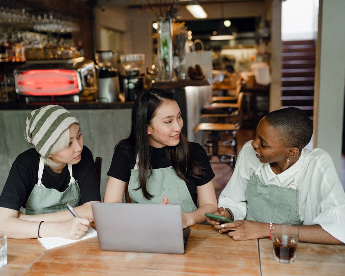 Three cafe staff in aprons working out the week together over a laptop
